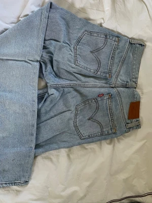 Levi's 501 ljusblå raka jeans - Klassiska Levi's 501 jeans i ljusblå denim med raka ben och hög midja. Jeansen har fem fickor, knappgylf och den ikoniska läderlappen bak i midjan. Snygga kontrastsömmar och den röda Levi's-taggen på bakfickan. Perfekta för en avslappnad och trendig look.
