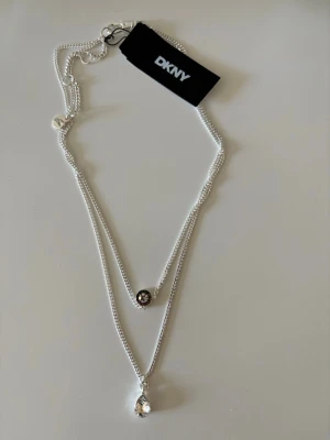 DKNY dubbelt halsband i silver - Snyggt dubbelt halsband från DKNY i silverfärg med två tunna kedjor. Ena kedjan har en rund pärldetalj med glittrande stenar och den andra har en droppformad klar sten som hänge. Fick i present men passar inte mig. Den har alla lappar kvar och kommer med originalförpackning! 