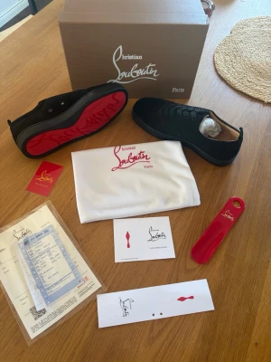 Svarta Christian Louboutin sneakers - Snygga svarta sneakers från Christian Louboutin med den ikoniska röda sulan. Skorna har en klassisk låg siluett, svarta skosnören och är tillverkade i slätt läder. Kommer med originalkartong, dustbag och tillbehör. Perfekta för dig som vill sticka ut med exklusiv stil.