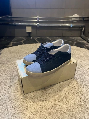 Blåa Golden Goose Superstar sneakers - Snygga Golden Goose Superstar sneakers i mocka med mörkblå och ljusblå paneler, vita detaljer och klassisk stjärnlogga på sidan. Skorna har vita sulor och mörkblå skosnören. Perfekta för dig som gillar en stilren men ändå unik look. Säljer då jag har tröttnat och söker nytt!