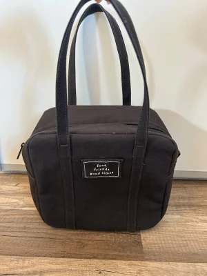 Mörkgrå kylväska med tryck från H&M Home  - Mörkgrå fyrkantig kylväska med dubbla handtag och dragkedja upptill från H&M Home. På framsidan finns ett tygmärke med texten 'food friends good times'. Måtten är 19 cm X 12 cm X 20 cm. Yttertyg: 100% bomull. Foder: 100% polyeten. Bård: 100% polyester. 
