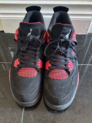 Svarta och röda Air Jordan 4 sneakers - Säljer ett par Air Jordan 4 sneakers i svart och rött med klassisk Jumpman-logga på plösen. Skorna har meshdetaljer, plastdetaljer vid snörningen och en vadderad insida. Ovandelen är i mocka och textil, och sulan är i gummi. Riktigt snygga och ikoniska sneakers för dig som gillar streetwear.