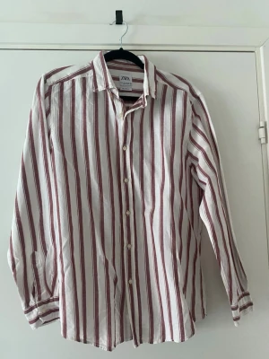 Randig skjorta från Zara i linne - Säljer en vit skjorta från Zara med vinröda ränder och relaxed fit. Skjortan har klassisk krage, knäppning framtill och långa ärmar. Tillverkad i luftigt linne, perfekt för en avslappnad stil.