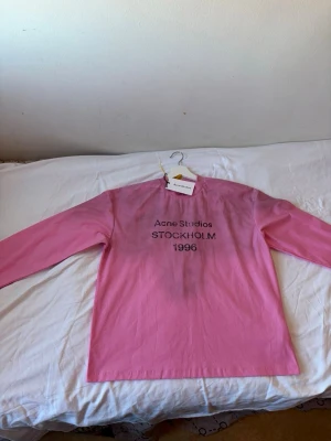 Rosa långärmad topp Acne Studios - Trendig rosa långärmad topp från Acne Studios med svart tryck framtill där det står 'Acne Studios STOCKHOLM 1996'. Rak passform och rund halsringning. Perfekt för dig som gillar stilrena och coola plagg.