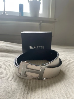Vändbart bälte med H-spänne - Snyggt vändbart bälte från Personal Tailor med en stor H-formad spänne i silver. Ena sidan är vit och den andra svart, vilket gör det enkelt att matcha till olika outfits. Tillverkat i skinn och levereras i originalförpackning.