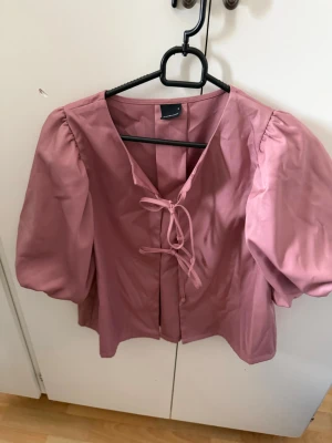 Rosa puffärmblus från Gina Tricot - Söt rosa blus från Gina Tricot med puffiga korta ärmar och knytning framtill. Blusen har en rak passform och är tillverkad i ett slätt tyg som ger en stilren look. Perfekt för att lyfta vilken outfit som helst.