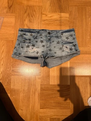 Jeansshorts med stjärnor och slitningar - Snygga ljusblå, lågmidjade jeansshorts med mörkblå stjärnor och slitna detaljer framtill. Superfina i superbra skick! Mått finns på sista bilden