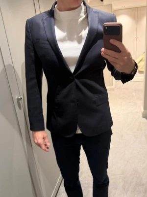 Mörk blå kostym i ull från J.Lindeberg  -  Säljer en stilren mörkblå kostym i ull med slimmad passform och snygga detaljer som knappar på ärmsluten och två framfickor. Kavajen har en bröstficka och är tillverkad i ett material med struktur som ger ett elegant intryck. Perfekt för dig som vill ha en snygg o tidlös look. Modell: Hopper 
