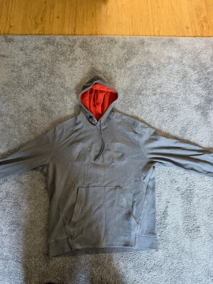 Grå hoodie från BOSS med röd huva - Snygg grå hoodie från BOSS med broderad logga på bröstet och röd insida i huvan. Klassisk känguruficka framtill och dragsko i huvan. Perfekt för en avslappnad och trendig stil.