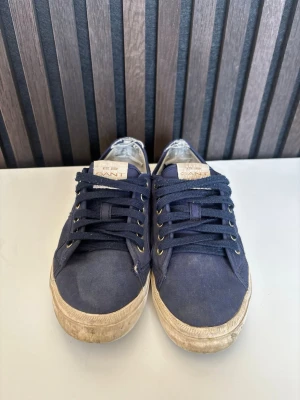 Marinblå sneakers från Gant - Snygga marinblå sneakers från Gant med klassisk låg profil och vita slitna sulor. Skorna har textilovandel, mörkblå snören och metallfärgade öljetter. Gant-logga på sidan och etikett på plösen. Perfekta för en avslappnad streetstyle.