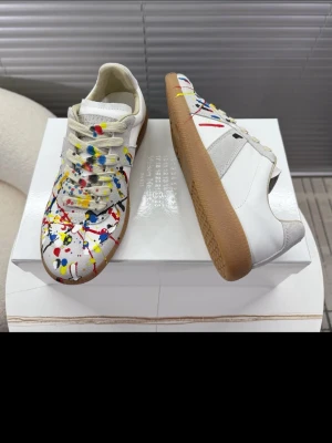 Maison Margiela sneakers med färgstänk - Unika sneakers från Maison Margiela i vitt läder och mocka med färgstänk i rött, blått, gult, grönt och svart. Klassisk låg modell med beige gummisula och vita snören. Perfekt för dig som vill sticka ut med en kreativ och trendig look.