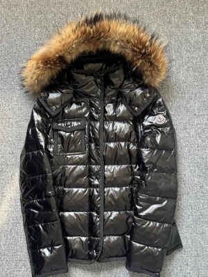 Svart glansig dunjacka Moncler - Svart glansig dunjacka från Moncler med avtagbar huva och stor pälskant. Jackan har en bröstficka med lock, två sidofickor med dragkedja och Moncler-logga på ärmen. Klassisk quiltad design och varm stoppning, perfekt för kalla vinterdagar.
