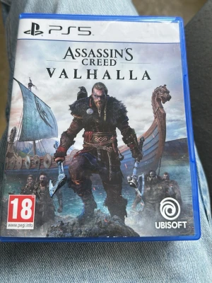 Assassin's Creed Valhalla PS5 - Assassin's Creed Valhalla till PS5 är ett actionäventyrsspel där du spelar som en viking. Omslaget har en blå plastbox med vikingatema, skepp och stridsyxor. Spelet är PEGI 18 och erbjuder episka räder, brutal närstrid och en öppen värld att utforska.
