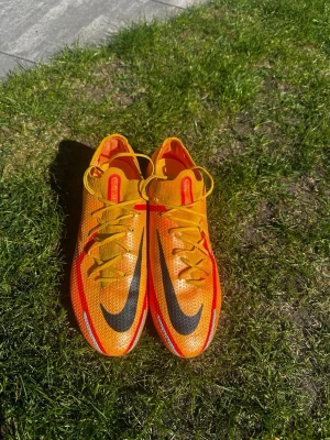 Nike Phantom GT2 orange fotbollsskor - Säljer ett par Nike Phantom GT2 fotbollsskor i en riktigt fet orange färg med röda och svarta detaljer. Skorna har snörning, låg profil och är tillverkade i syntetmaterial med textur för bättre bollkontroll. Perfekta för dig som vill sticka ut på planen.