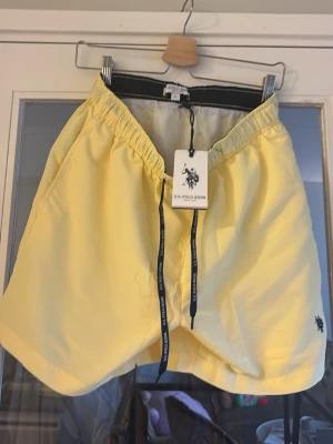 Gula badbyxor från U.S. Polo Assn. Helt nya med tag  - Helt nya med tag!!!  Snygga gula badbyxor från U.S. Polo Assn. med svart logga broderad på ena benet och svarta snören med märkets namn. Klassisk modell med resår i midjan och innerbyxa i mesh. Perfekta för stranden eller poolen.