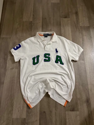 Vit pikétröja från Polo Ralph Lauren - Vit pikétröja med grönt och blått 'USA'-tryck på bröstet och blå logga från Polo Ralph Lauren. Kortärmad modell med klassisk krage och orange detaljer i sidorna. Siffra 3 i blått på ena ärmen. Perfekt för en sportig och stilren look.