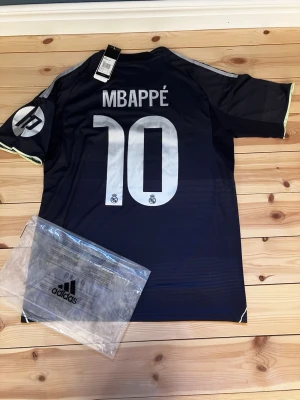 Real Madrid Mbappé matchtröja Adidas - Snygg mörkblå Real Madrid matchtröja från Adidas med Mbappé och nummer 10 på ryggen. Tröjan har vita detaljer, klubbmärke och sponsortryck framtill samt HP-logga på ärmen. Tillverkad i lätt och ventilerande material, perfekt för fotbollsträning.