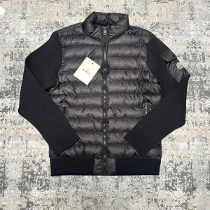 Svart Cardigan - Stilren svart cardigan med quiltad framsida och släta svarta ärmar. Västen har hög krage, dragkedja framtill och logga på ärmen. Materialet är en kombination av dun och textil, vilket ger en modern look.