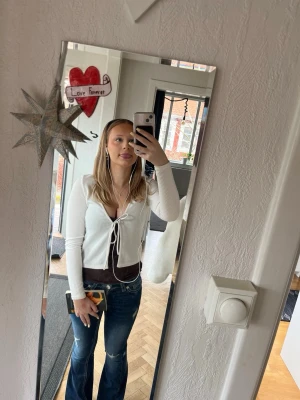 Vit ribbad långärmad topp H&M - Trendig vit ribbad topp från H&M med lång ärm och knytning framtill. Djup v-ringning och croppad passform som ger en snygg siluett. Perfekt att styla med jeans eller kjol för en cool look.