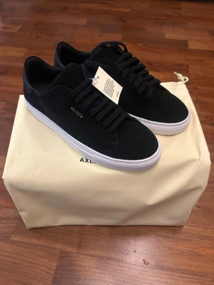 NYA Svarta sneakers från Axel Arigato - Helt nya Axel Arigato sneakers köpta från NK. Kan visa recept som bevis. De var för små så jag kan inte returnera dem så jag säljer dem på Plick för rabatt.  Snygga svarta sneakers från Axel Arigato med mocka ovandel och klassiska svarta snören. Skorna har en vit, platt sula och diskret guldlogotyp på sidan. Perfekt för en ren och modern look.