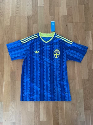 Blå Sverige fotbollströja Adidas - Säljer en blå Sverige fotbollströja från Adidas med gula detaljer och mönstrade ränder. Tröjan har korta ärmar, V-ringning med gul kant och Sveriges landslagsemblem på bröstet. Perfekt för dig som vill visa ditt stöd för landslaget.