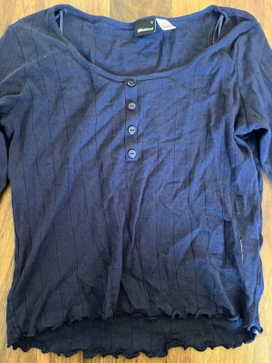 Marinblå topp med knappar Gina Tricot - Mörkblå topp från Gina Tricot med rund halsringning och fyra knappar framtill. Toppen har lång ärm, diskret hålmönster och vågig kant nertill. Perfekt för en trendig och avslappnad look, använd 1 gång.
