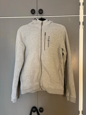 Ljusgrå hoodie  - Ljusgrå hoodie  med dragkedja framtill och en bröstficka med zip. Tillverkad i mjukt sweatshirtmaterial och har en broderad logga på bröstet. Klassisk passform med ribbade muddar och huva för extra komfort.