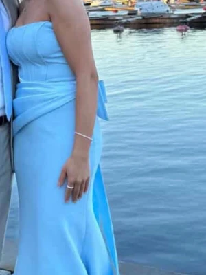 Ljusblå axelbandslös långklänning - Säljer en elegant ljusblå axelbandslös långklänning med figurnära passform och draperade detaljer i midjan. Klänningen har en slät yta och en stilren look, perfekt för speciella tillfällen. Använd en kväll och kemtvättad