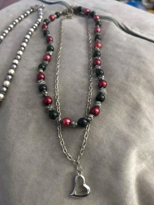 Dubbelhalsband med pärlor och hjärthänge - Snyggt dubbelhalsband med silverfärgad kedja och pärlor i vinrött och svart. Ena kedjan har ett öppet hjärthänge i metall och den andra är dekorerad med pärlor och små detaljer i silverfärg. Perfekt accessoar för att lyfta din outfit.