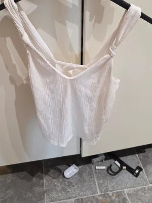 Somrig vit topp med knutdetaljer – H&M (XS) - Säkra den perfekta bastoppen för sommaren! Denna vita dröm har en fantastisk våfflad struktur i tyget som ger den både liv och karaktär. De snygga knutdetaljerna på axelbanden ger en extra feminin och unik touch. ✨  Tack vare blandningen av viskos och elastan faller toppen otroligt fint och är superstretchig och bekväm att bära. Perfekt till ett par höga jeans eller en blommig kjol!  