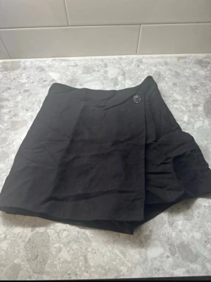 Svart skort med knappdetalj - Stilren svart skort med omlott-look framtill och en snygg knappdetalj i midjan. Skorten har shorts under och kjol över, vilket ger en trendig och bekväm känsla. Perfekt för dig som gillar en enkel men cool stil. Materialet känns mjukt och lätt.