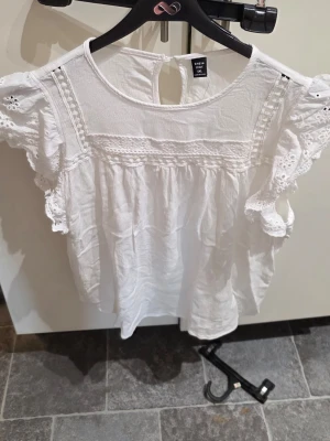 Vit blus med spets från SHEIN Curve - Söt vit blus från SHEIN Curve med broderade spetsdetaljer och volangärmar. Blusen har rund halsringning, knapp i nacken och är tillverkad i ett lätt och luftigt tyg. Perfekt för en fräsch och somrig look.