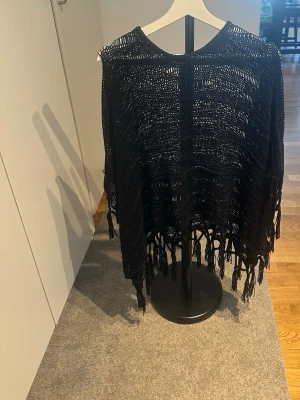 Svart poncho med fransar - Säljer en svart poncho med snygga fransar längs kanten. Ponchon har ett öppet, luftigt mönster och den passar till många olika outfits. Maskor har gått upp på vissa ställen.