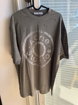 Grå Acne Studios t-shirt med tryck - Cool grå t-shirt från Acne Studios med stort vitt logotryck framtill. Oversized passform och korta ärmar. T-shirten är i mjuk bomull och har en avslappnad vibe med små slitningar för en trendig look.