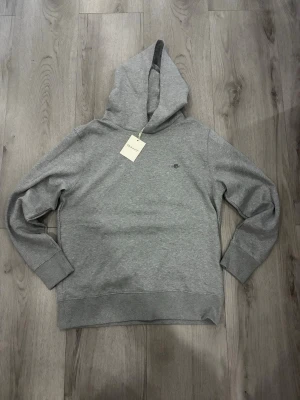Grå hoodie från Gant med broderad logga - Snygg gråmelerad hoodie från Gant med liten broderad logga på bröstet. Klassisk modell med huva och ribbade muddar vid ärmslut och nederkant. Perfekt för en avslappnad och stilren look.