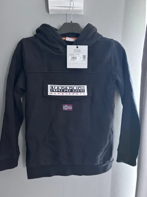 Svart hoodie från Napapijri - Svart hoodie från Napapijri med stor logotyp och norsk flagga på bröstet. Klassisk huva och ribbade muddar. Perfekt för en avslappnad och cool stil. Tillverkad i mjukt material som känns skönt mot huden. Storlek 162, 12-14 år 