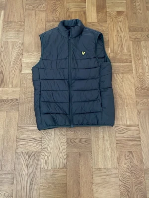 Skogsgrön dunväst från Lyle & Scott - Snygg dunväst från Lyle & Scott med gul logga på bröstet. Västen har ståkrage, dragkedja framtill och quiltad design. Knappt använd och nypris är 500kr