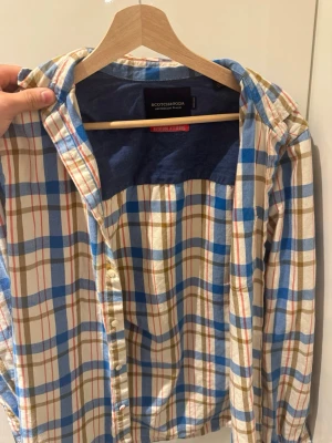 Rutig skjorta från Scotch & Soda - Rutig skjorta från Scotch & Soda i blått, vitt och beige med inslag av rött. Skjortan har klassisk krage, knäppning framtill och långa ärmar. Tillverkad i mjuk bomull och passar perfekt för en avslappnad stil.
