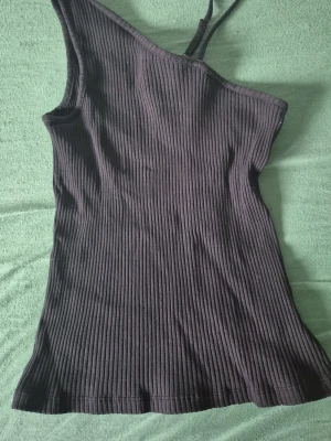 Svart ribbad oneshoulder topp - Snygg svart ribbad topp med oneshoulder-design och smal axelrem. Perfekt för dig som vill ha en trendig och stilren look. Toppen är figurnära och passar till många olika outfits.