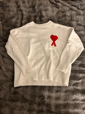 Vit stickad tröja med rött hjärta - Vit stickad tröja från AMI Paris med ett stort rött hjärta och bokstaven A broderat på bröstet. Tröjan har rund hals, långa ärmar och breda ribbade muddar vid ärmslut och nederkant. Perfekt för dig som gillar stilrena detaljer.