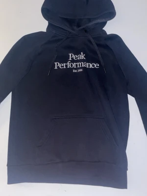 Svart hoodie från Peak Performance - Svart hoodie från Peak Performance med vit logga och text 'Est. 1986' på bröstet. Klassisk känguruficka framtill, dragsko i huvan och mjukt material på insidan. Perfekt för en avslappnad och sportig stil.