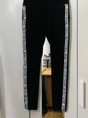 Svarta leggings med logga från Illusion - Svarta leggings med vita sidoränder där det står 'ILLUSION' längs hela benet. Tight passform och elastiskt material som sitter snyggt. Perfekta för en sportig och trendig look. Vet inte varför men den har inte storlek på men jag skulle gissa på att den passar bra för 11-15 åringar 💕