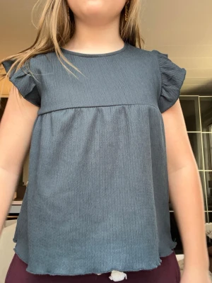 Mörkblå blus med volangärmar - Supersöt mörkblå blus med korta volangärmar och rund hals. Blusen har en lätt rynkad struktur och är tillverkad i ett mjukt, tunt material som ger ett luftigt intryck. Perfekt för dig som gillar en enkel men ändå söt stil.