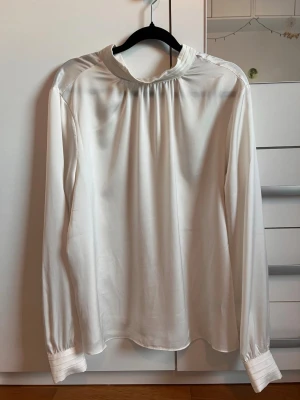Vit satinblus från H&M - Stilren vit blus i satin från H&M med hög krage och långa ärmar. Blusen har diskreta rynkdetaljer vid halsen och breda manschetter vid ärmsluten. Perfekt för en chic och elegant look.