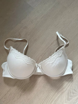 Vit vadderad bh med spetsdetalj - Säljer en vit vadderad bh från H&M med bygel och justerbara axelband. Bh:n har fina spetsdetaljer längs kupkanten och är tillverkad i ett ribbat tyg som ger en snygg struktur. Perfekt för dig som vill ha en enkel men ändå söt bh.
