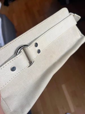 Beige kuvertväska i canvas - Stilren beige kuvertväska i canvas med detaljer i ljust konstläder och silverfärgade metalldelar. Väskan har en avlång form och en dekorativ rem på framsidan.  Lite fläckig. 