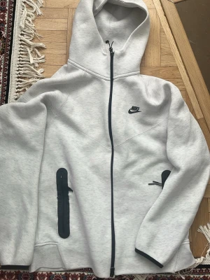 Ljusgrå Nike hoodie med dragkedja - Snygg ljusgrå hoodie från Nike med hel dragkedja framtill och svart Nike-logga på bröstet. Två svarta dragkedjeförsedda fickor på sidorna och justerbar huva. Tillverkad i mjukt sweatshirtmaterial, perfekt för chill eller streetwear.