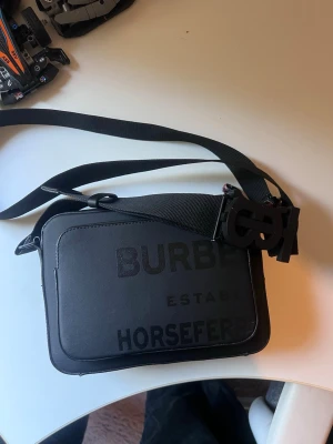 Burberry Axelväska  - Cool svart axelväska från Burberry med stor logga på framsidan och justerbar axelrem. Väskan har en unik spännesdetalj med B-formad låsning och är tillverkad i slitstarkt material. Perfekt för att bära dina viktigaste grejer med stil.