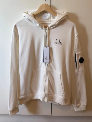 Vit hoodie från C.P. Company - Snygg vit hoodie från C.P. Company med dragkedja framtill, huva och fickor. Klassisk logga på bröstet och ikonisk linsdetalj på ärmen. Perfekt för en clean och stilren look.