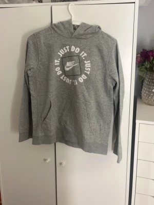 Grå Nike hoodie med tryck - Snygg grå hoodie från Nike med vit text 'Just Do It.' i cirkel och Nike-logga på bröstet. Klassisk känguruficka framtill och huva. Perfekt för en avslappnad och sportig stil. Mjuk bomullsblandning som är skön att ha på sig.